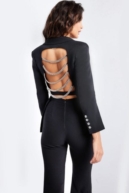 Model wearing Jovani 26144 black pantsuit with open back and chain detailing.