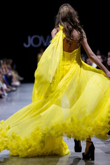 Model wearing Jovani S38104 yellow gown with open back and dramatic cape, back view.