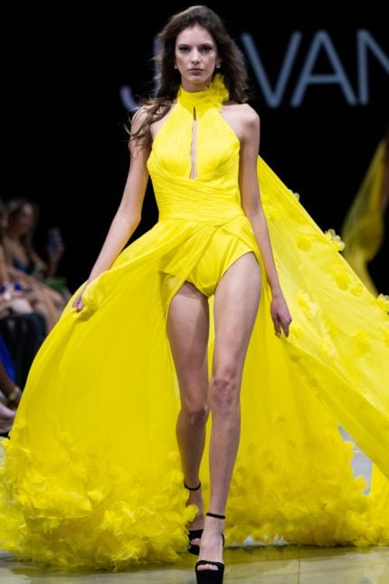 Model wearing Jovani S38104 yellow gown with halter neckline and flowing cape, front view.