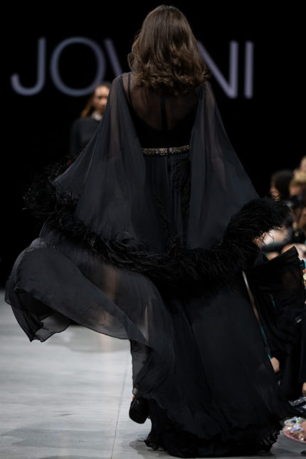 Model wearing S39276 black gown, back view with sheer feathered sleeves.