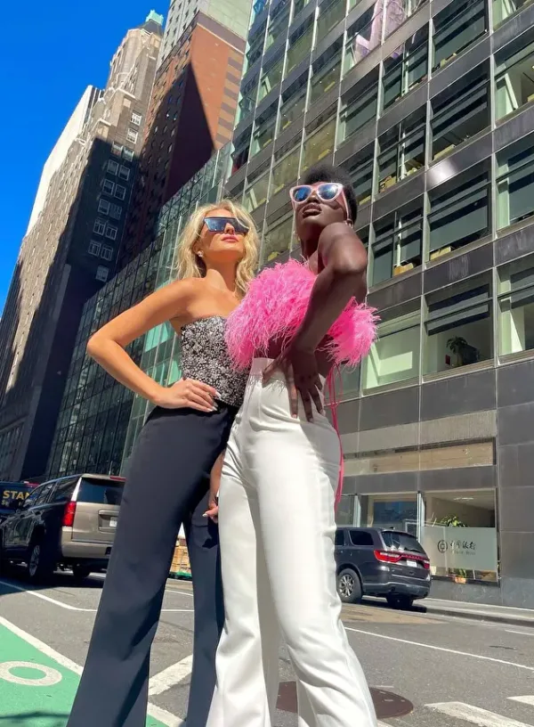 Two fashionable women standing confidently on a city street with tall buildings in the background. One woman is wearing a strapless top with black pants and sunglasses, while the other is wearing a pink feathered top with white pants and sunglasses.